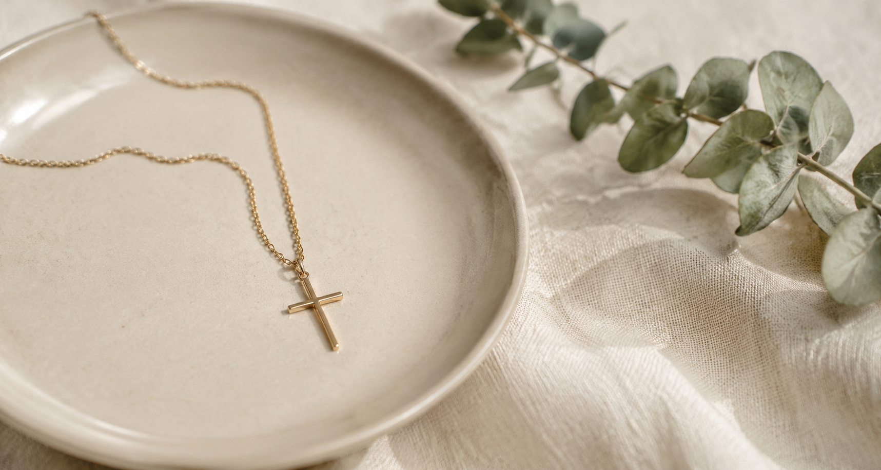 Gold cross necklace on warm cream ceramics and linen